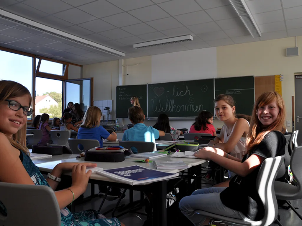 Zwei Mädchen drehen sich am Tisch zur Kamera um, an der Tafel steht „Willkommen“.